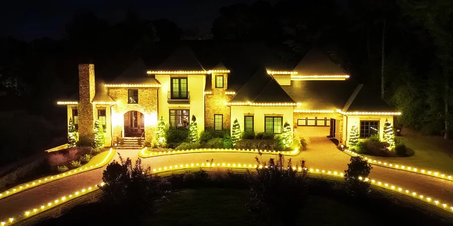 Large stone estate home with warm white roofline lighting and lit trees