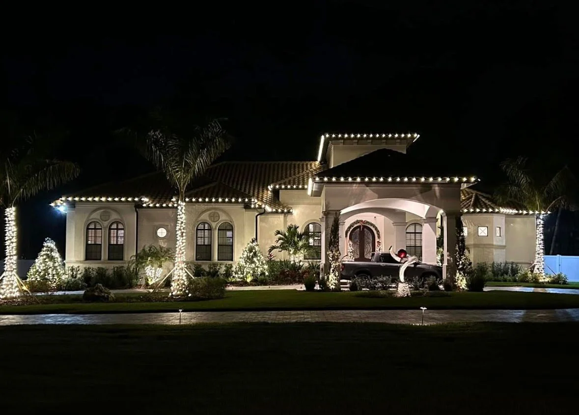 Single-story Spanish-style estate home with tile roof, warm white roofline lighting, and palm trees wrapped in lights