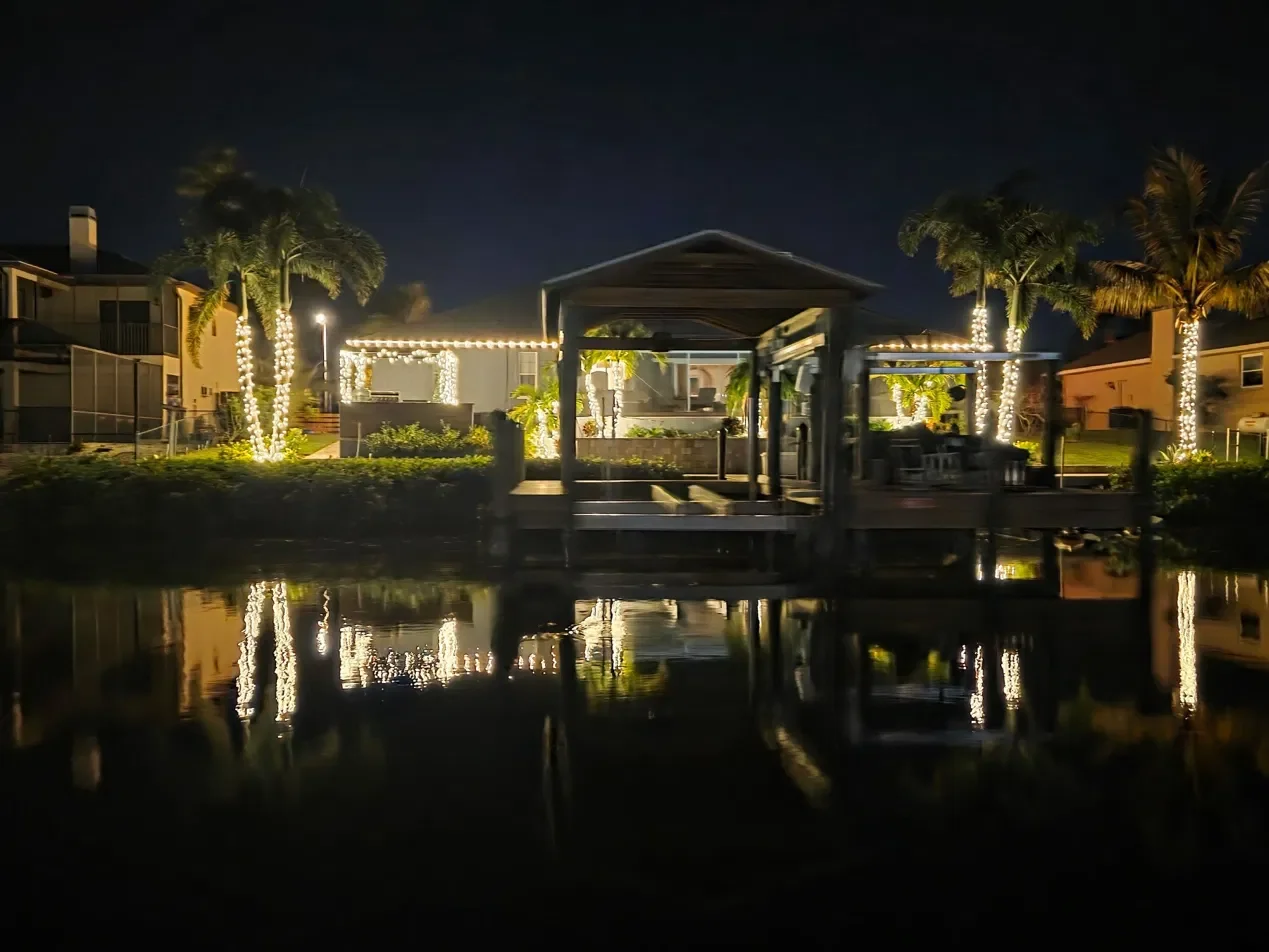 Waterfront home and dock with palm trees wrapped in warm white Christmas lights reflecting on the water