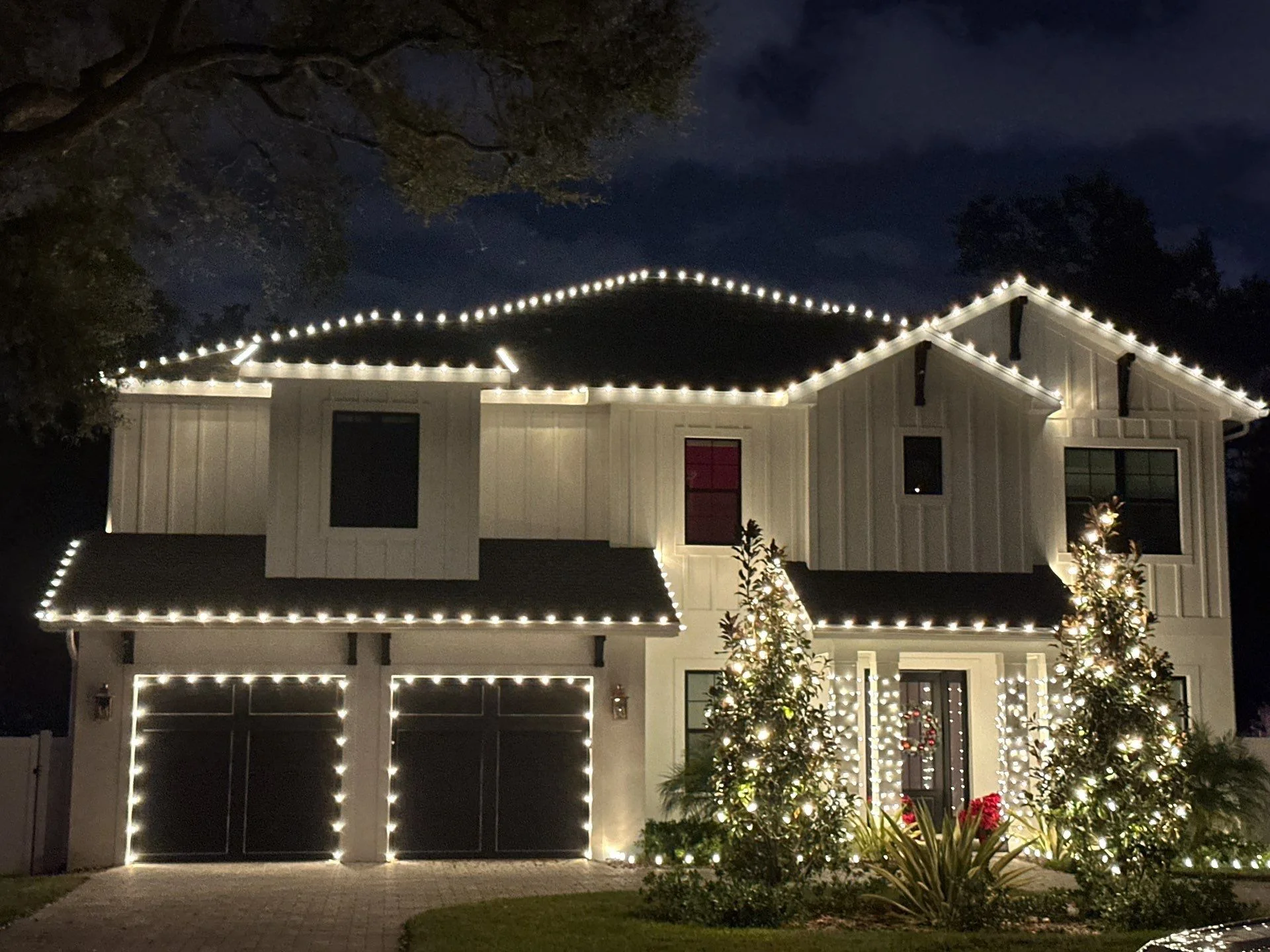 Two-story white farmhouse-style home with warm white roofline outline lighting and wrapped trees